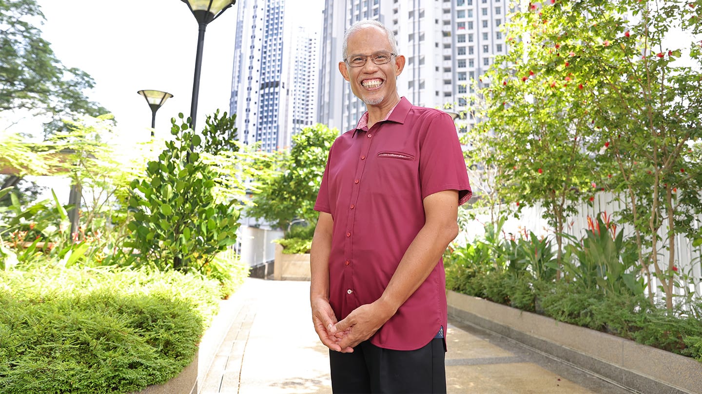 Man in glasses and red shirt, smiling outdoors with plants and tall buildings.